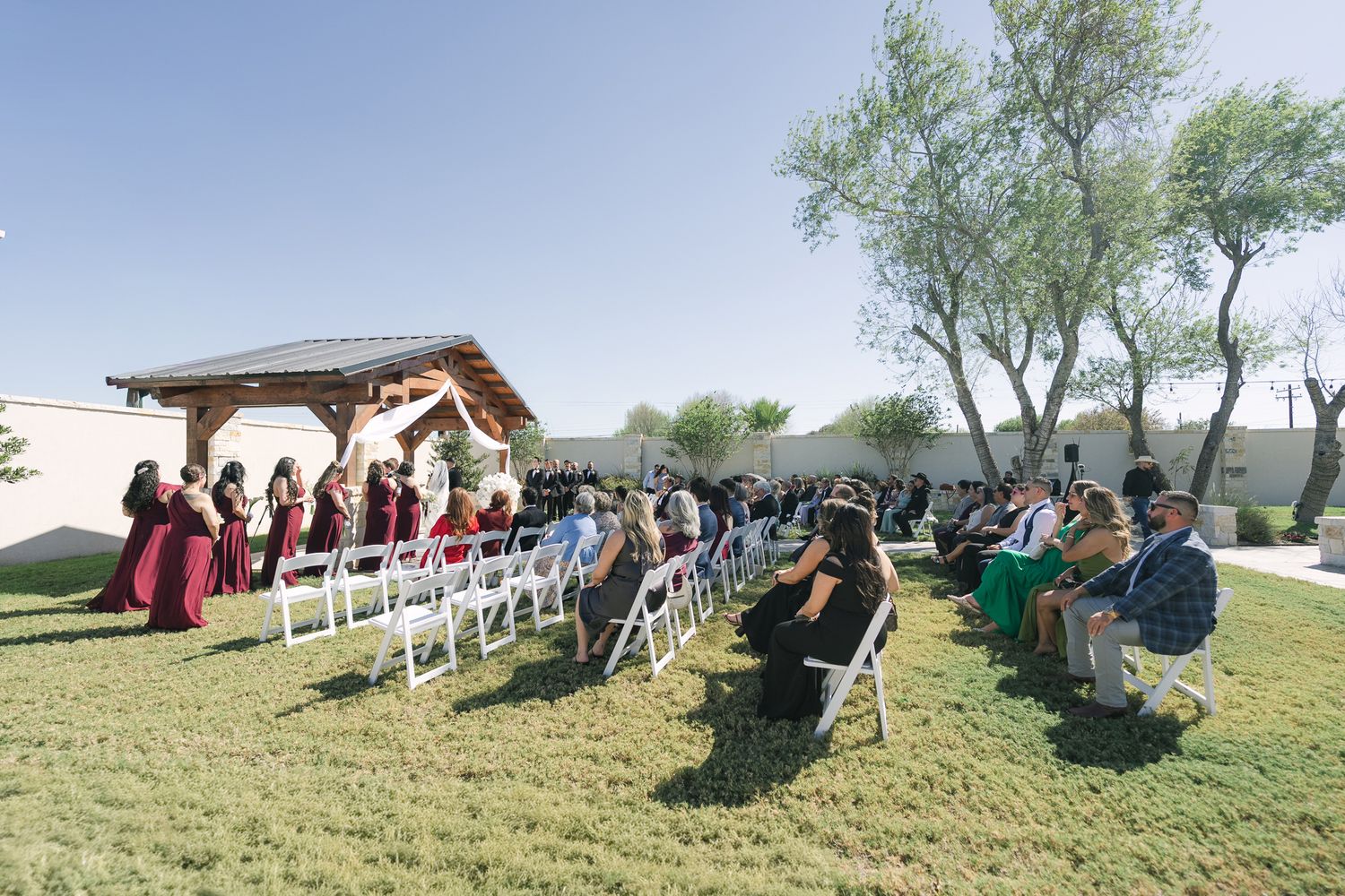 Outdoor wedding ceremony with guests seated on white chairs facing a wooden gazebo altar on a sunny day.