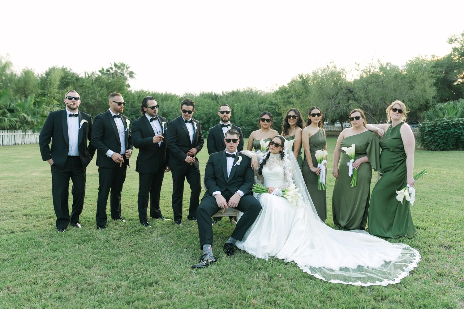 Wedding party posing outdoors, bride in white gown, groom in tuxedo, groomsmen in black suits, bridesmaids in green dresses.