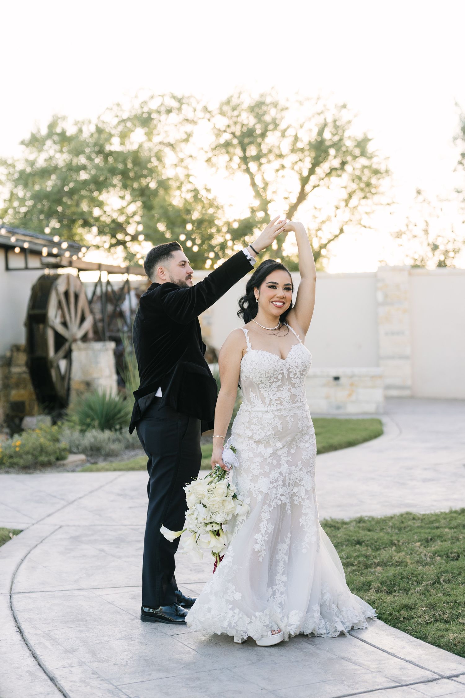 A groom in a black tuxedo twirls his bride in a floral wedding gown outdoors at golden hour.
