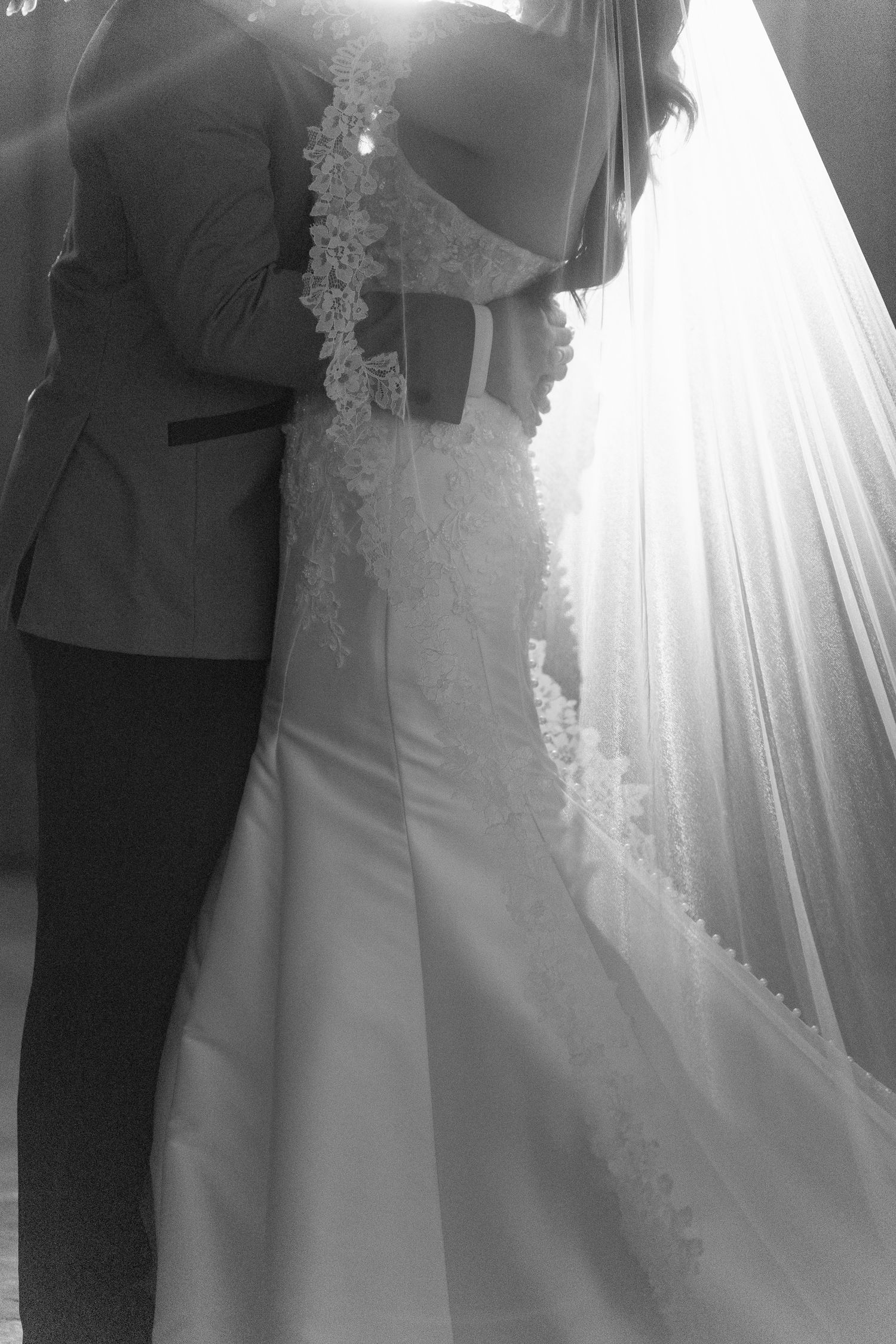 Black and white photo of bride and groom embracing, showcasing lace wedding dress and flowing veil in soft backlight.
