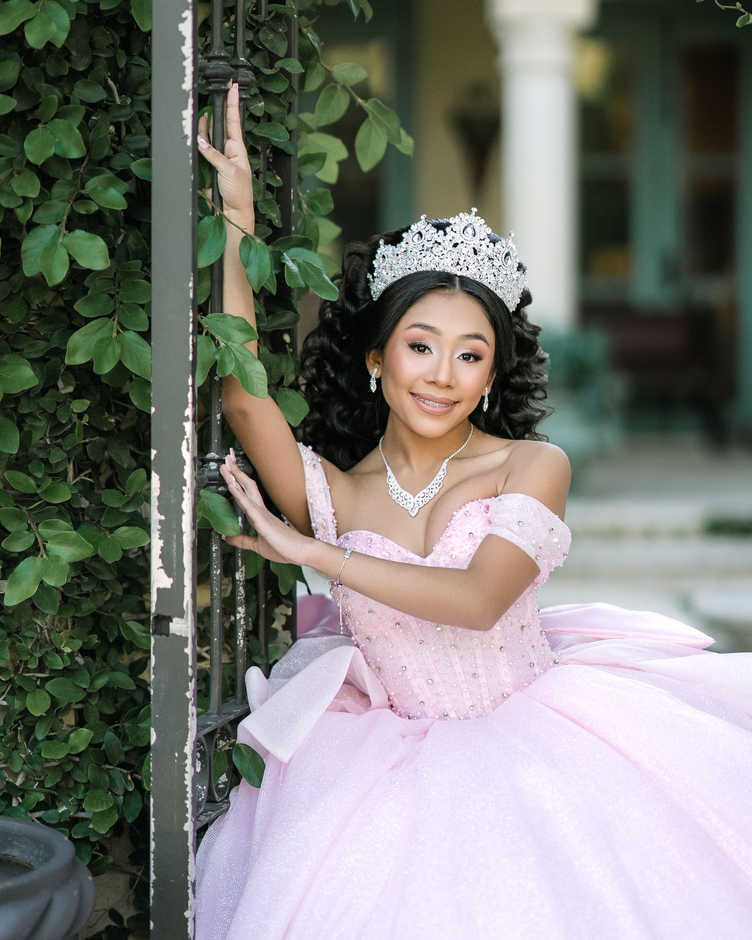 A person in a pink quinceañera dress and tiara poses elegantly by a vine-covered pillar.