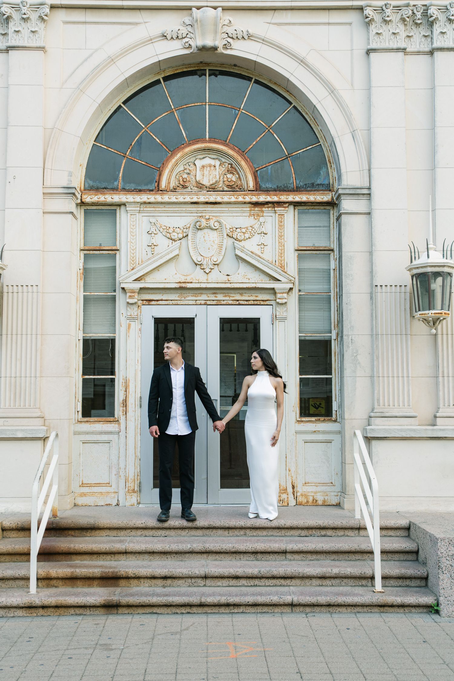 A couple in formal wear stands holding hands in front of an ornate white building entrance.