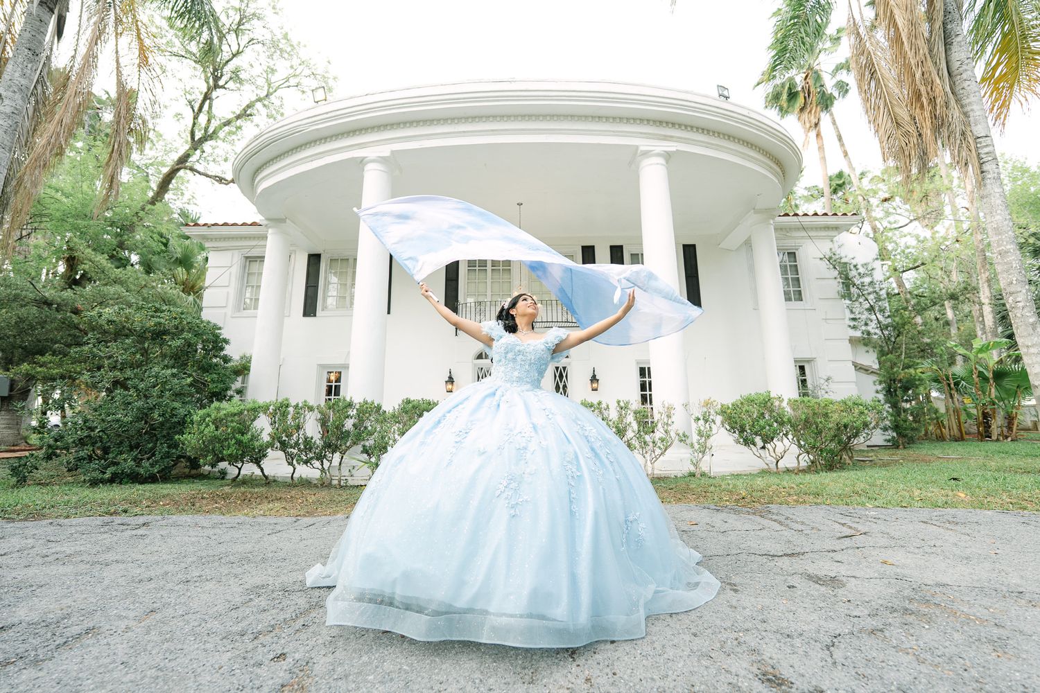 Flowing light blue ballgown photographed in outdoor garden setting with white architectural details.