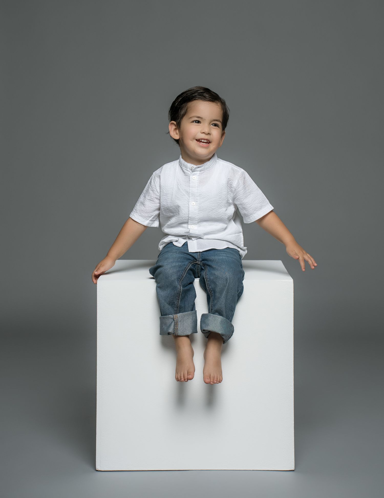 Child sitting barefoot on white cube wearing casual attire in studio setting.