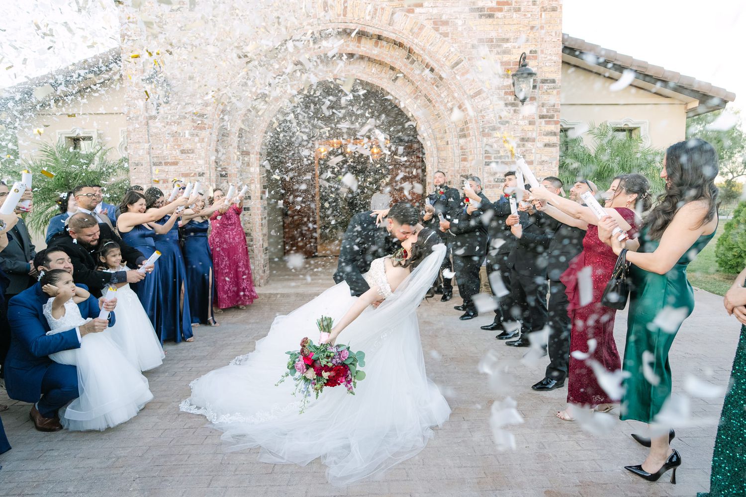Wedding couple celebrating under shower of confetti with guests outside stone archway.