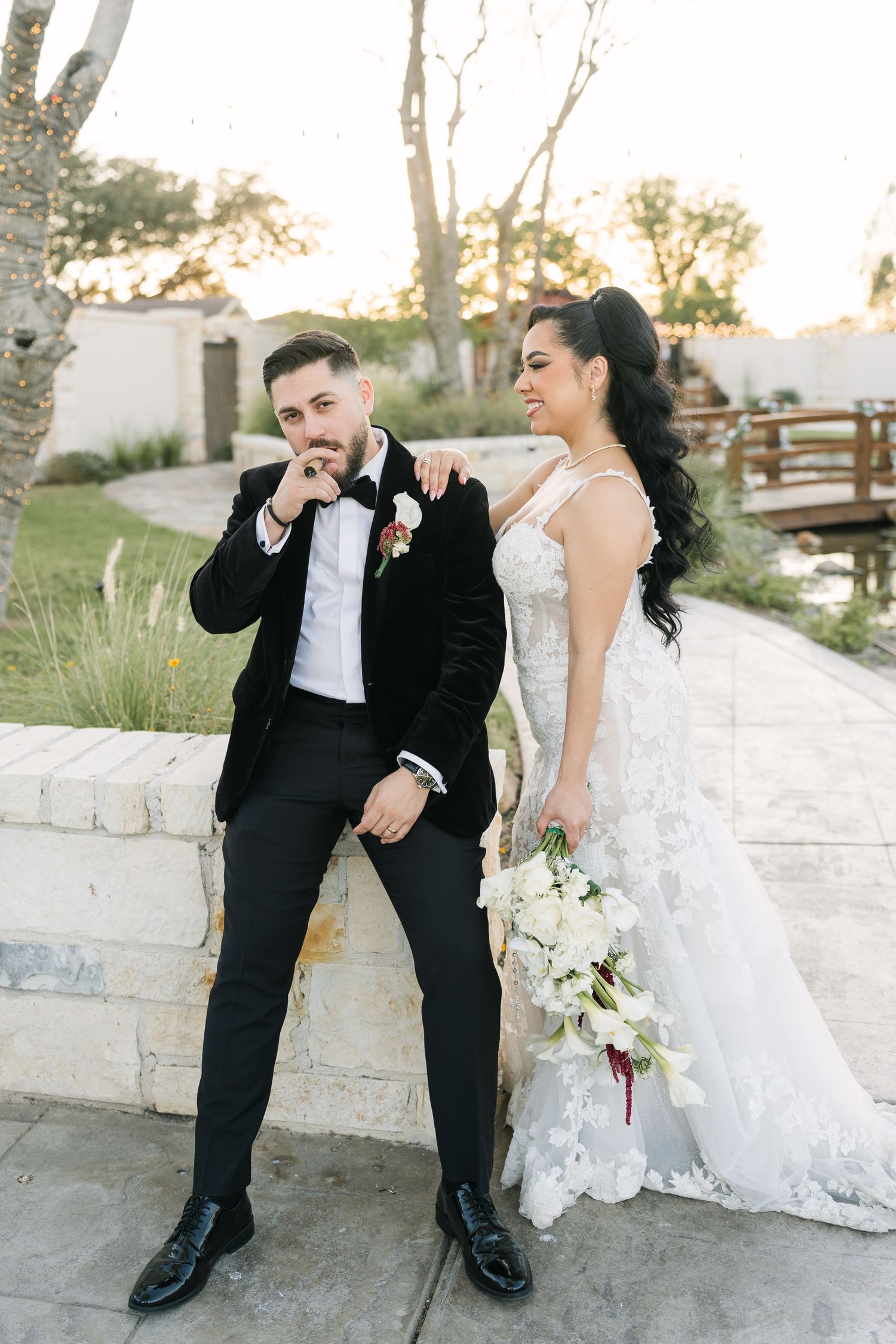 Bride in lace gown holding white bouquet stands with groom in black tuxedo kissing her hand at outdoor wedding venue.