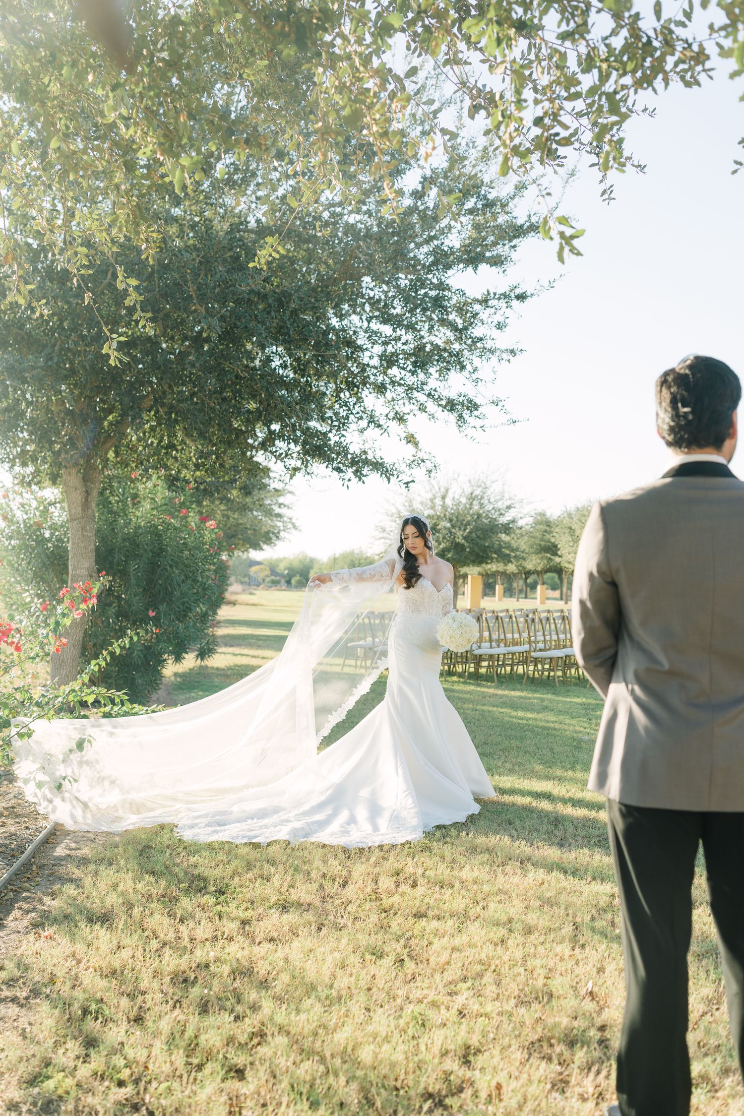 A bride and groom stand in a sunlit field with trees as the bride's veil flows dramatically in the breeze.