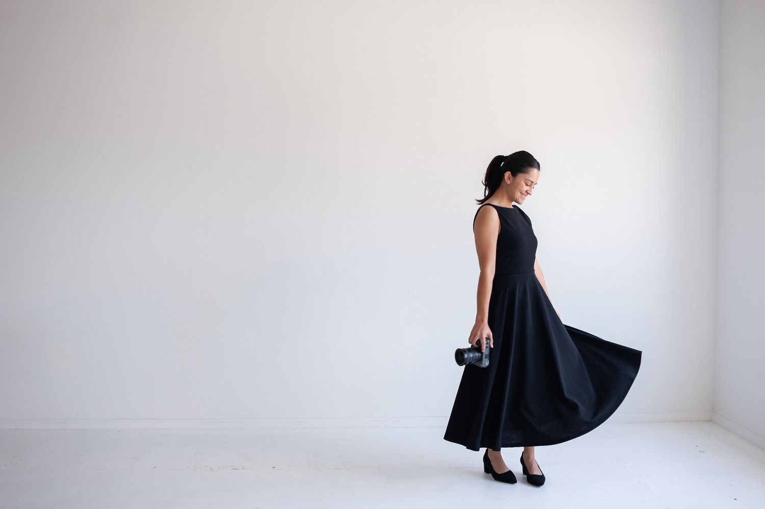 Woman in elegant black sleeveless midi dress holding clutch, posing gracefully in minimalist white studio.