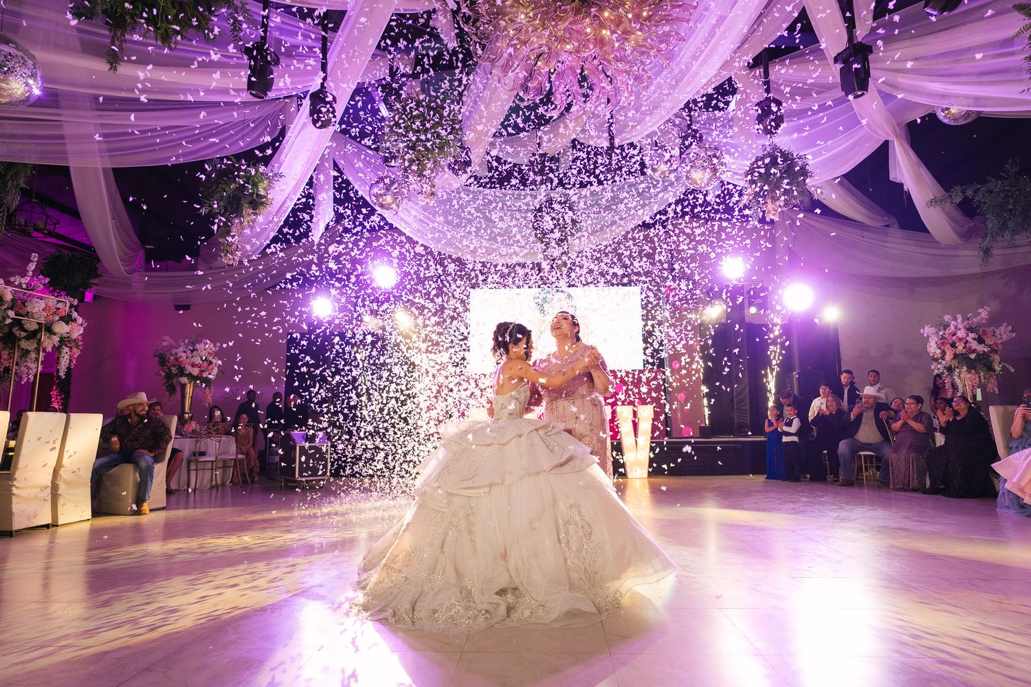 A wedding celebration with a grand dance floor illuminated by purple lights and falling confetti creates a magical atmosphere.