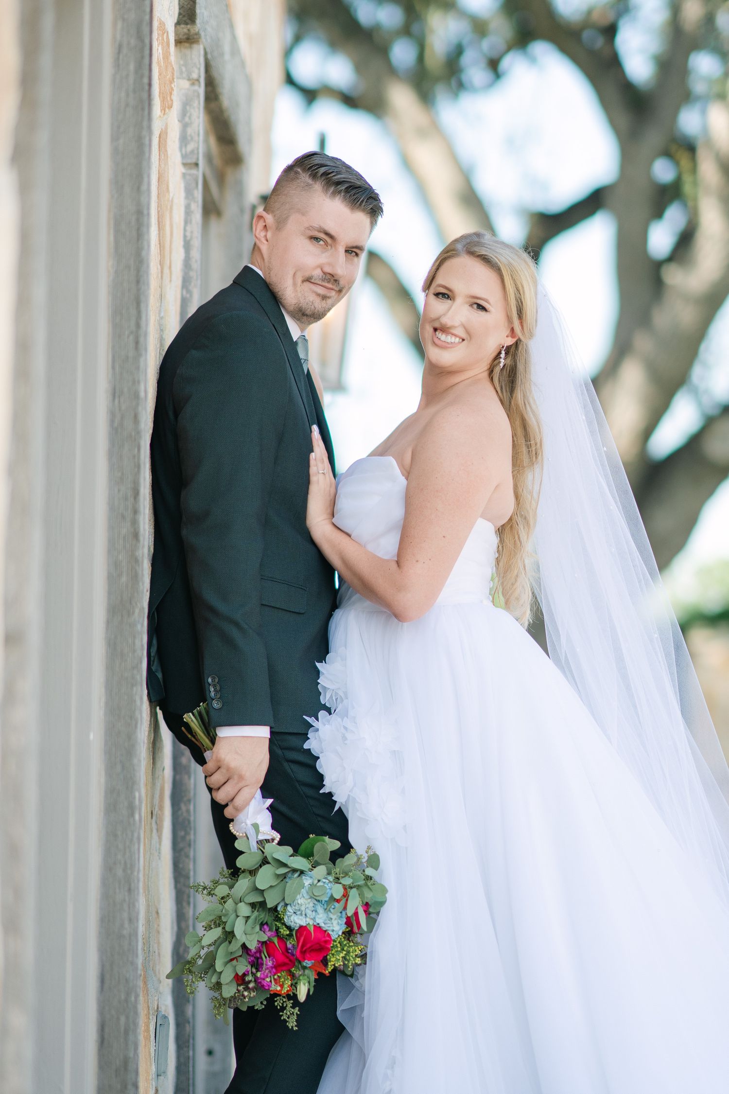 Wedding portrait of couple posed against stone wall with colorful floral bouquet.