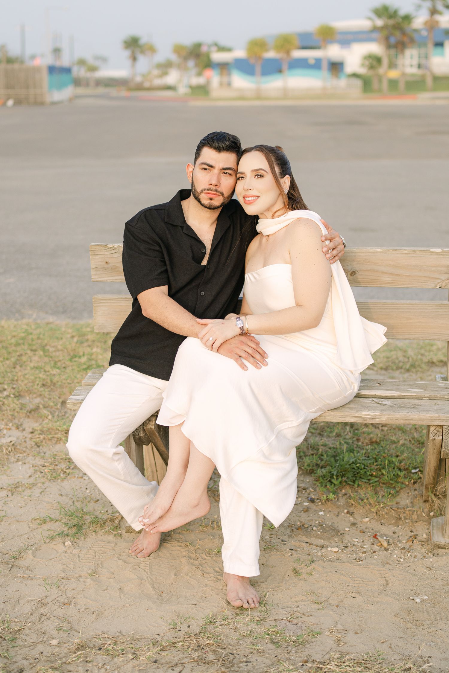 Couple in white and black attire sitting casually on a wooden bench near a beach.