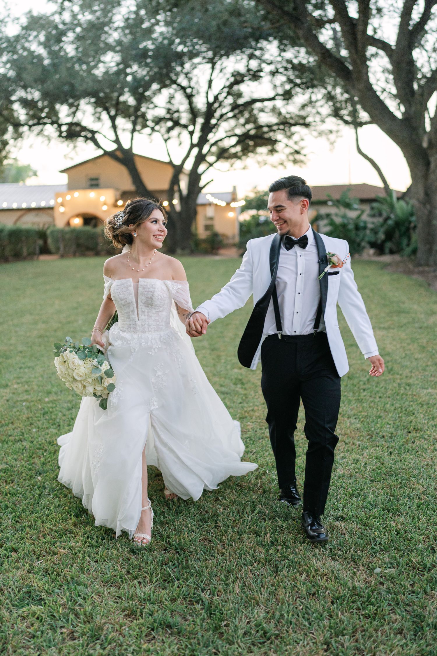A newlywed couple walks hand in hand across a grassy yard with trees in the background during their wedding day.