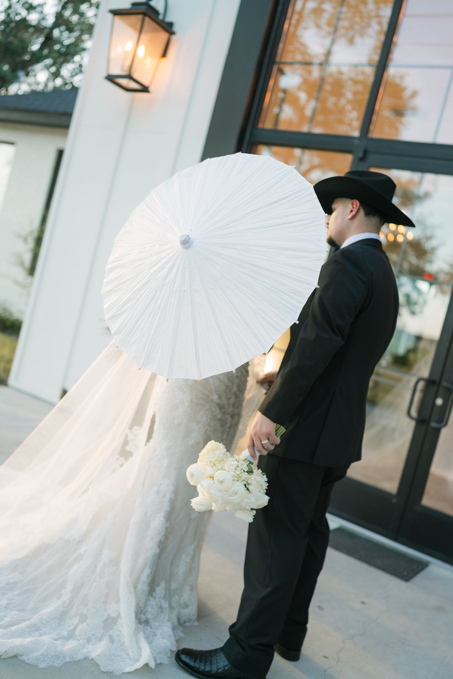 Wedding couple holding white parasol and bouquet, groom in black suit and cowboy hat, bride in lace gown outside venue.