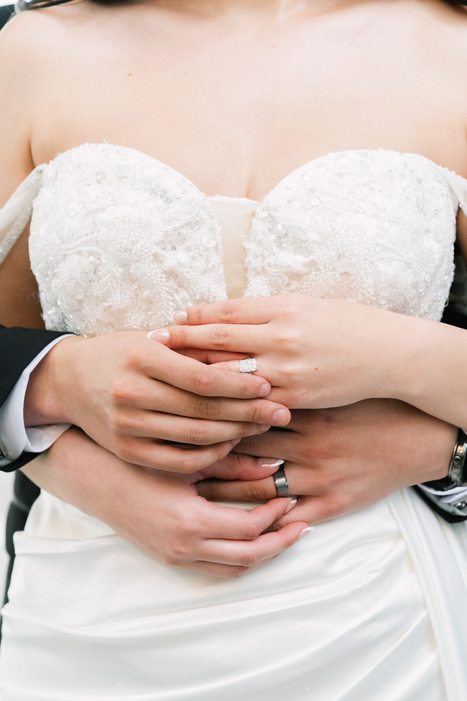 Close-up of bride and groom's hands embracing, showing wedding rings against a white lace dress.