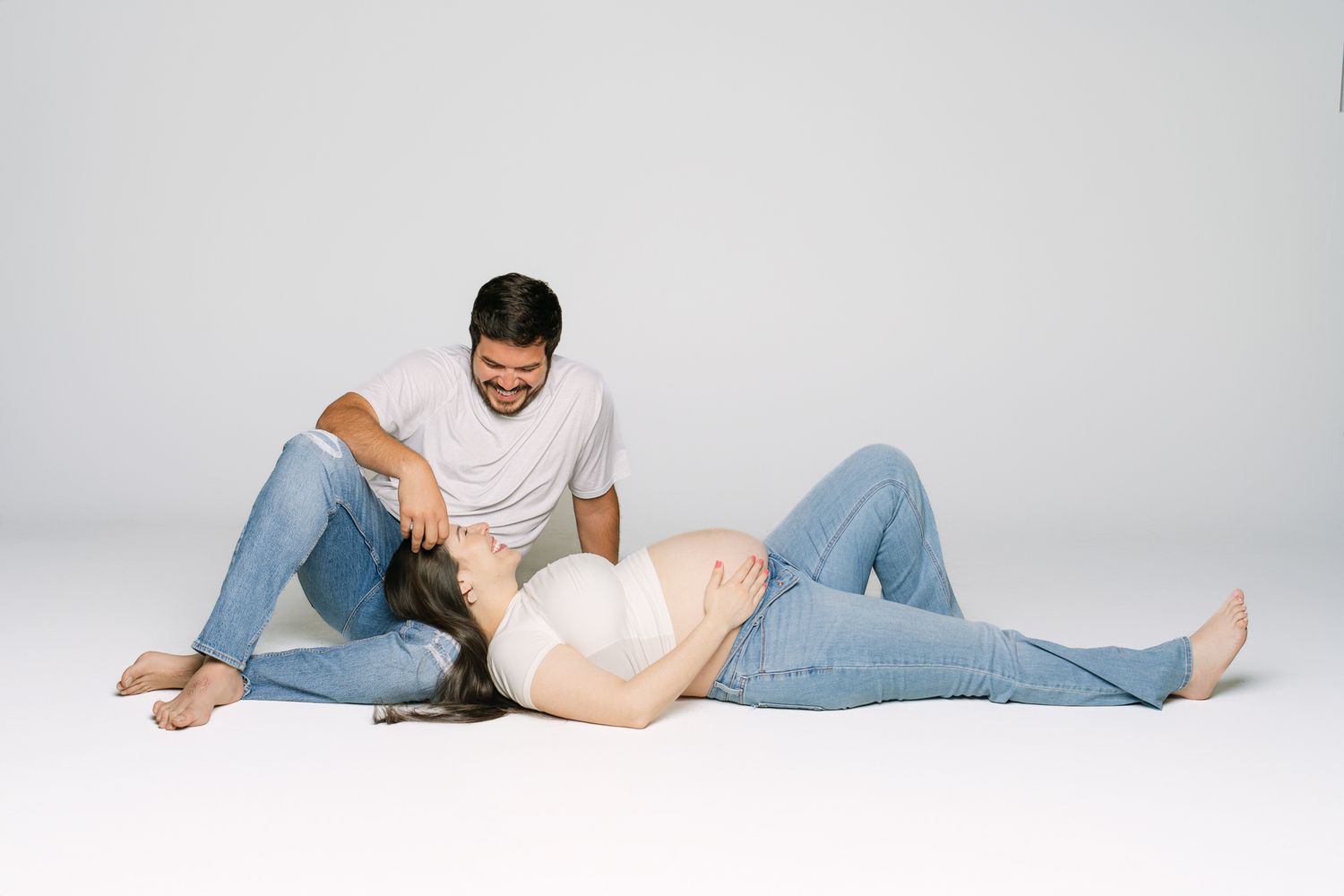 Couple resting together on white studio floor wearing casual jeans and white shirts.