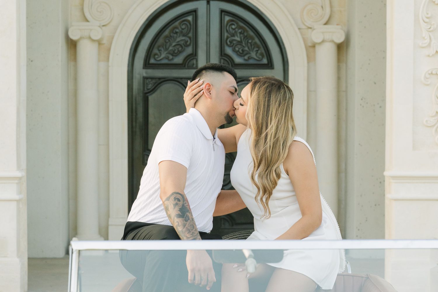 A couple in white outfits shares a tender kiss on a balcony.