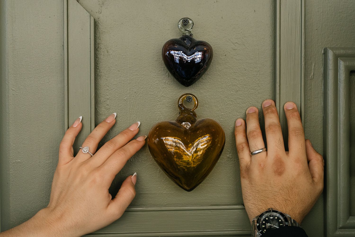 Two hands with rings reach toward decorative heart ornaments on a sage green door.