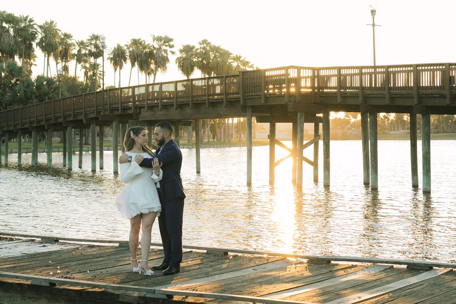 Bride and groom share a romantic kiss on a wooden dock at sunset with a bridge and palm trees in the background.
