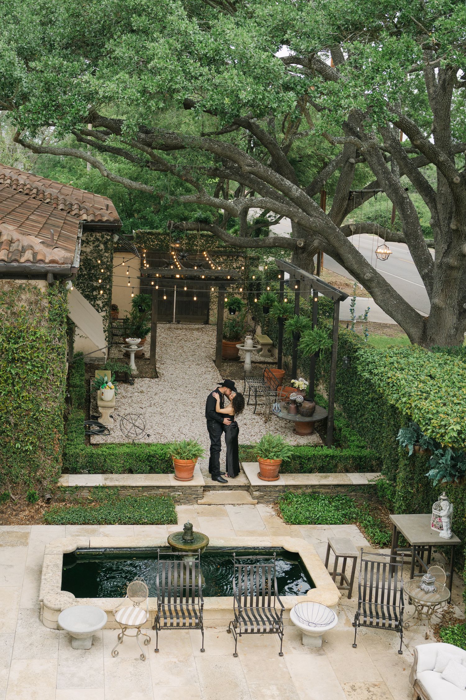 Couple sharing a romantic kiss in an elegant garden courtyard with a fountain, string lights, and lush greenery.