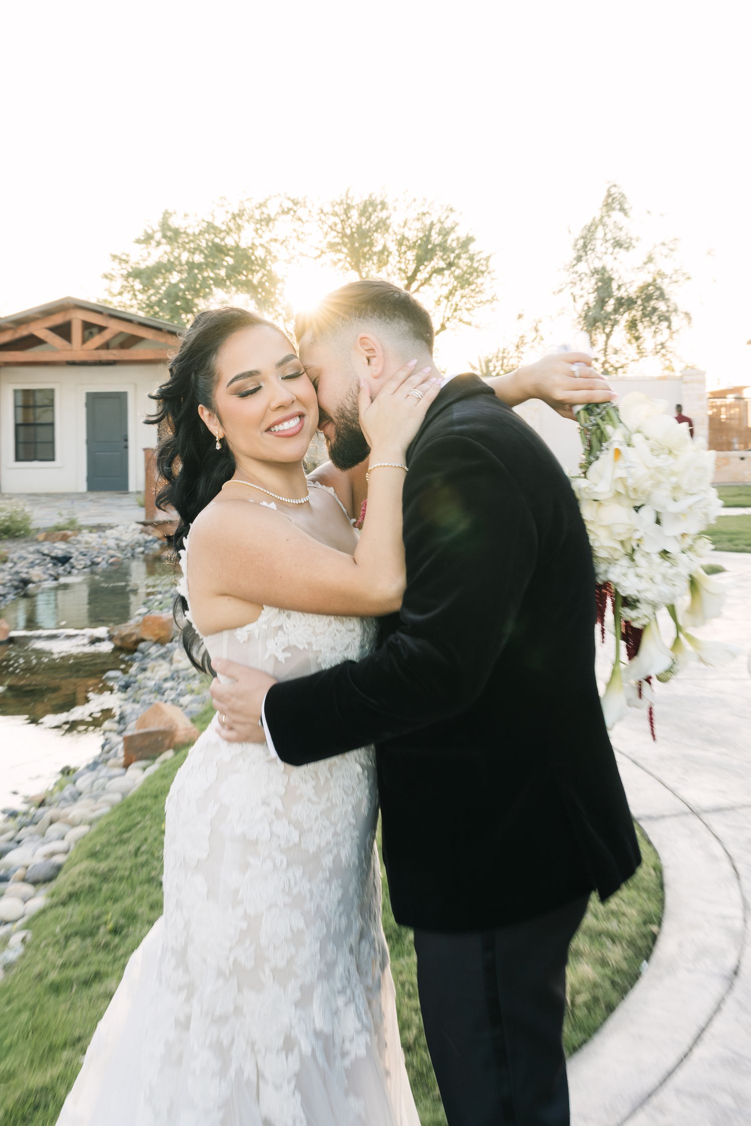 Bride in lace gown and groom in black tuxedo embrace at golden hour, with white bouquet and sunlit outdoor venue.