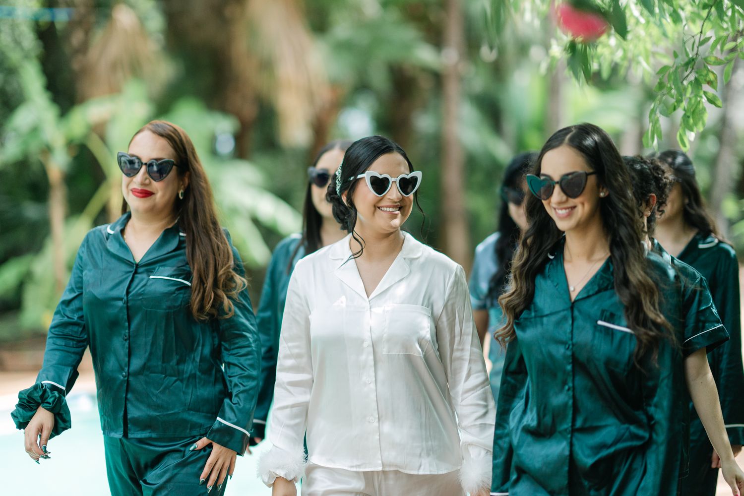 Bridal party wearing coordinated emerald green outfits and sunglasses walk together outdoors.