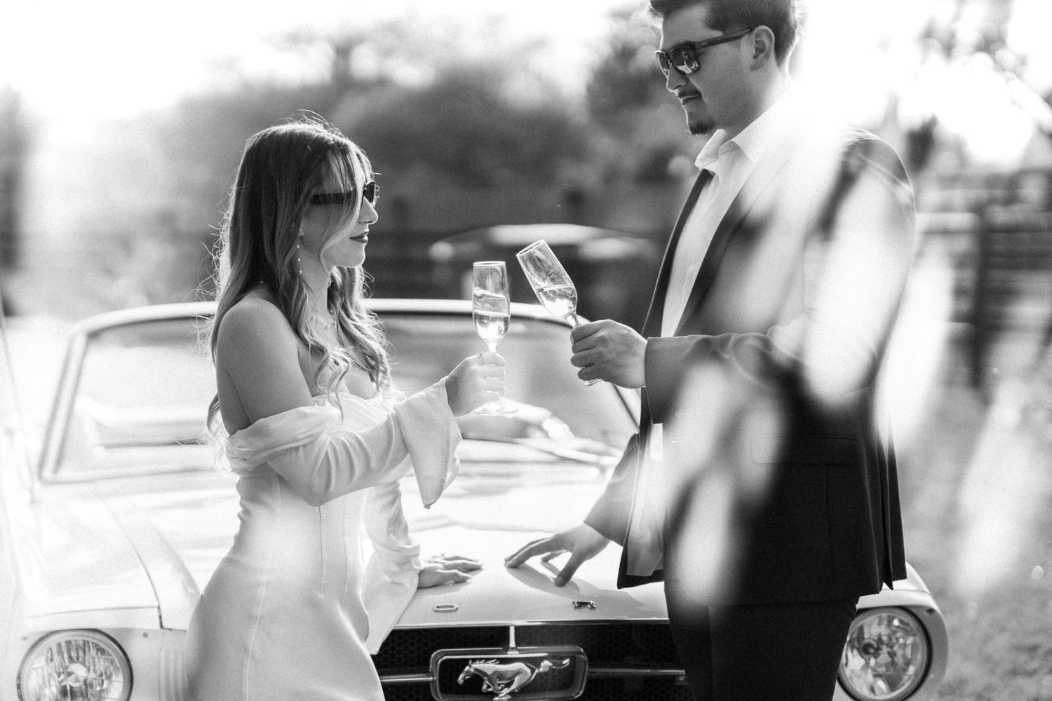 A black and white photo of a couple exchanging rings by a classic car.