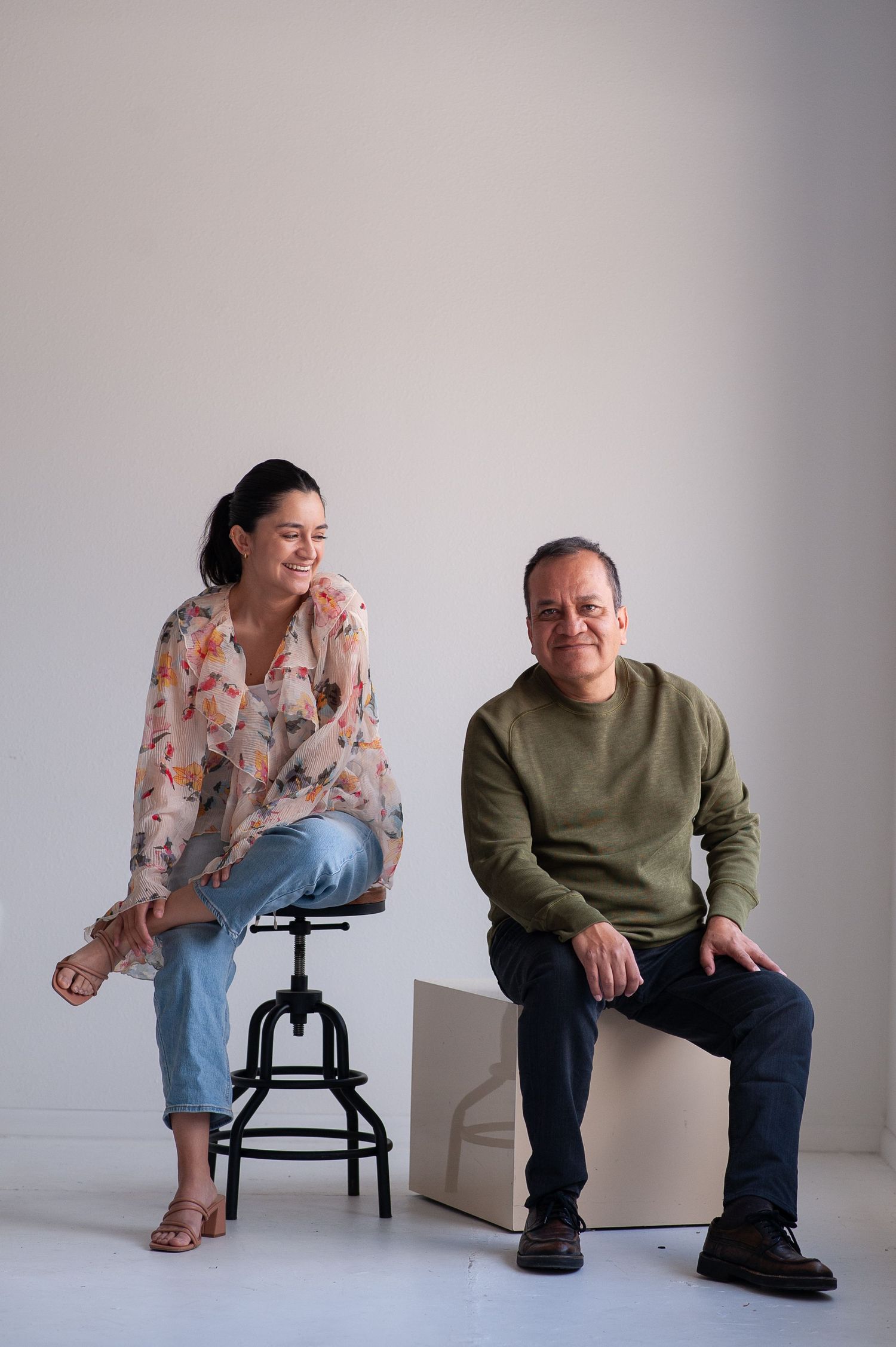 A woman in floral top and jeans sits on a stool beside a man in green sweater on a white box.