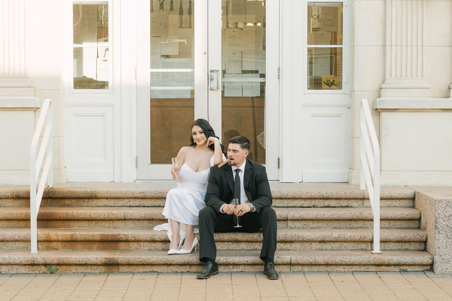 A couple in formal attire sits on white stone steps of a building entrance.