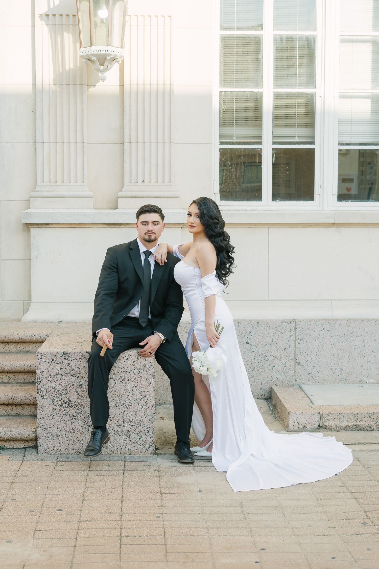 Wedding couple in formal attire posing creatively on stone steps outside a classical building.