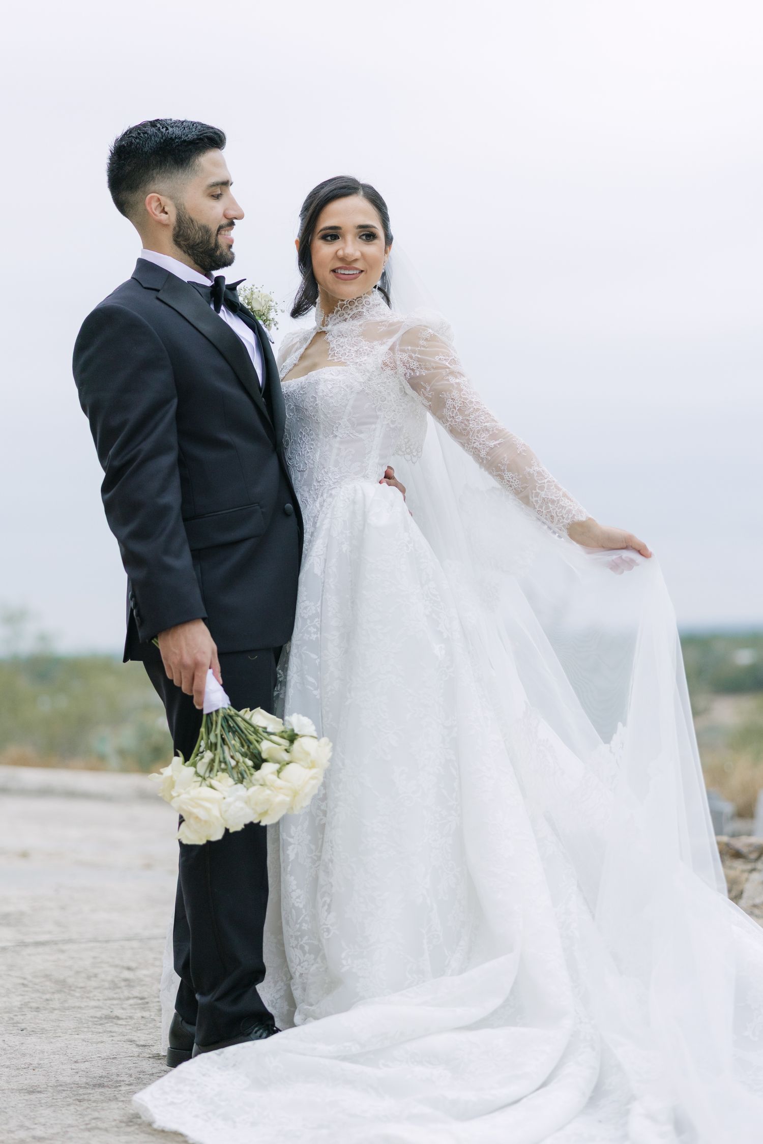 Bride in white lace gown and groom in black tuxedo pose together outdoors holding white roses bouquet.