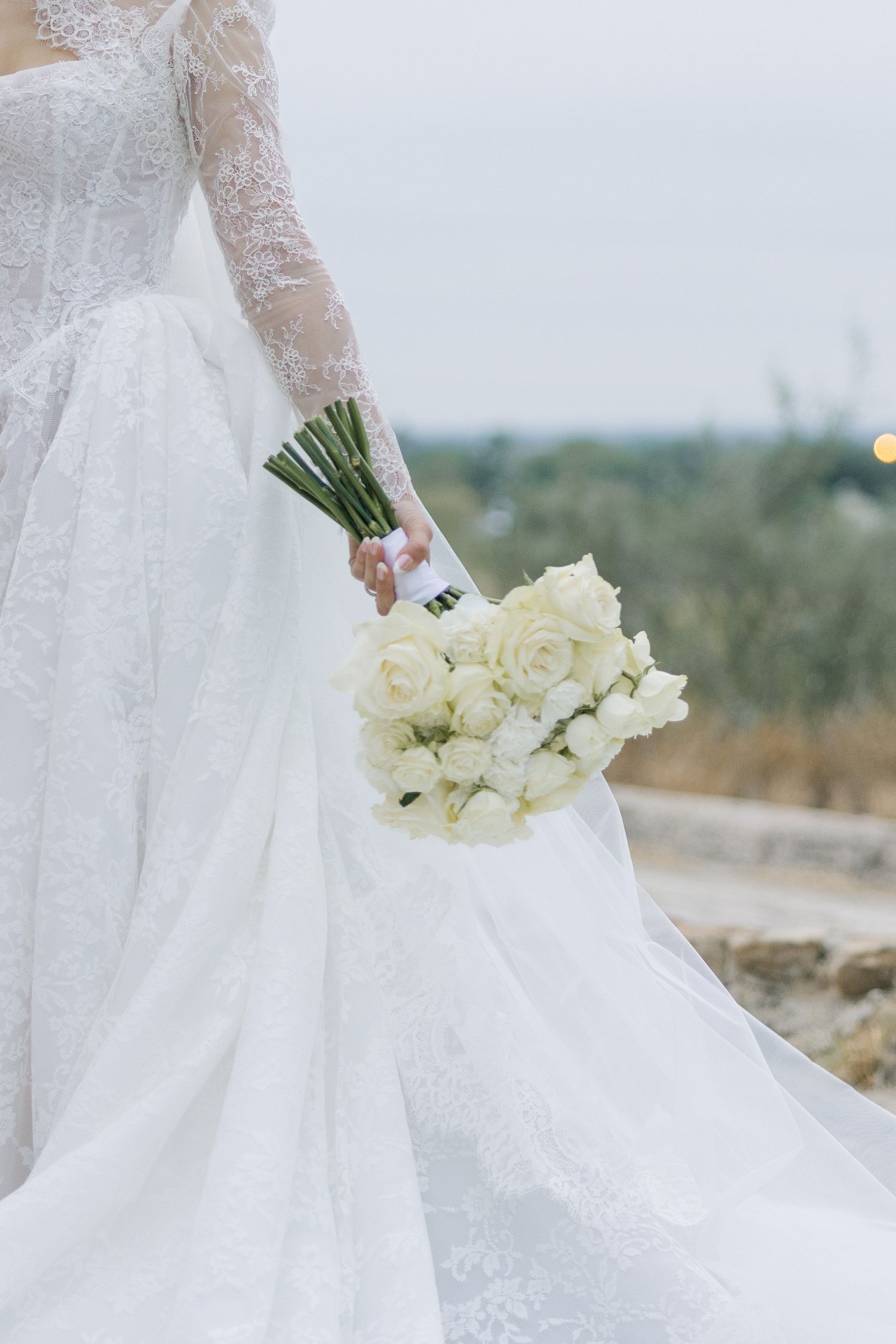 Bride in white lace wedding gown holding elegant bouquet of white roses outdoors.