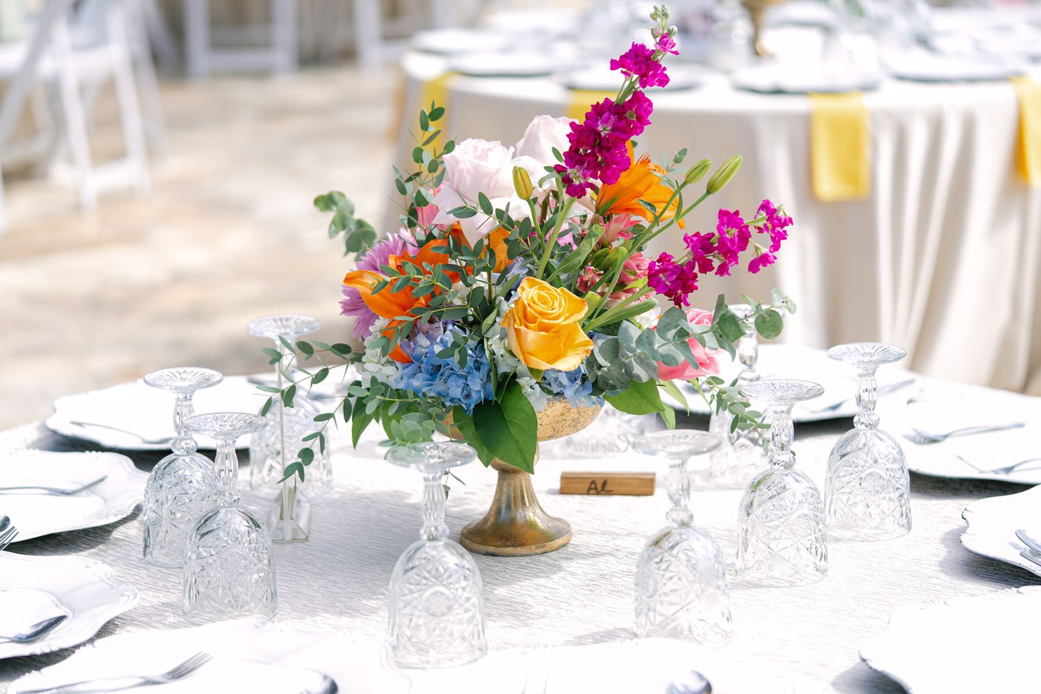 Elegant wedding reception centerpiece with bright florals in gold vase on round table.