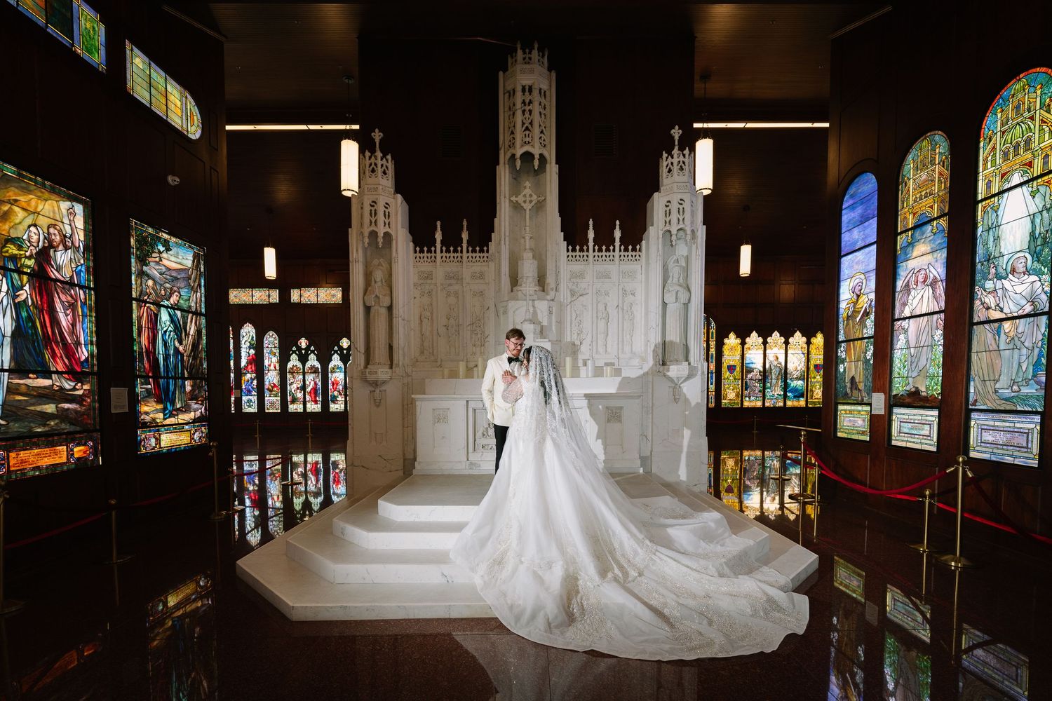 Bride and groom pose before a stunning white cathedral backdrop with colorful stained glass windows in an elegant wedding venue.