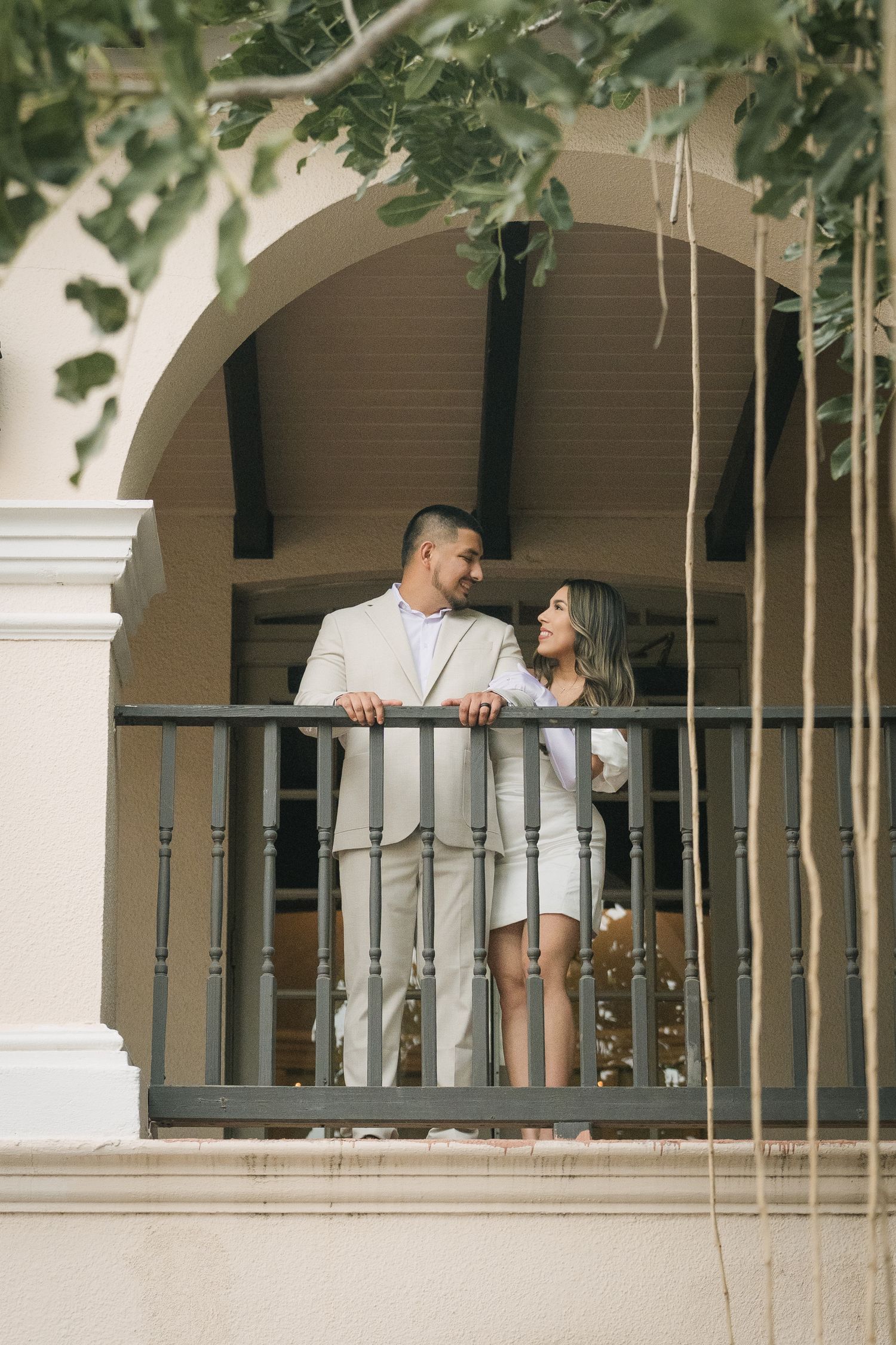 Couple in elegant attire sharing a romantic moment on a balcony framed by an arch and lush greenery.