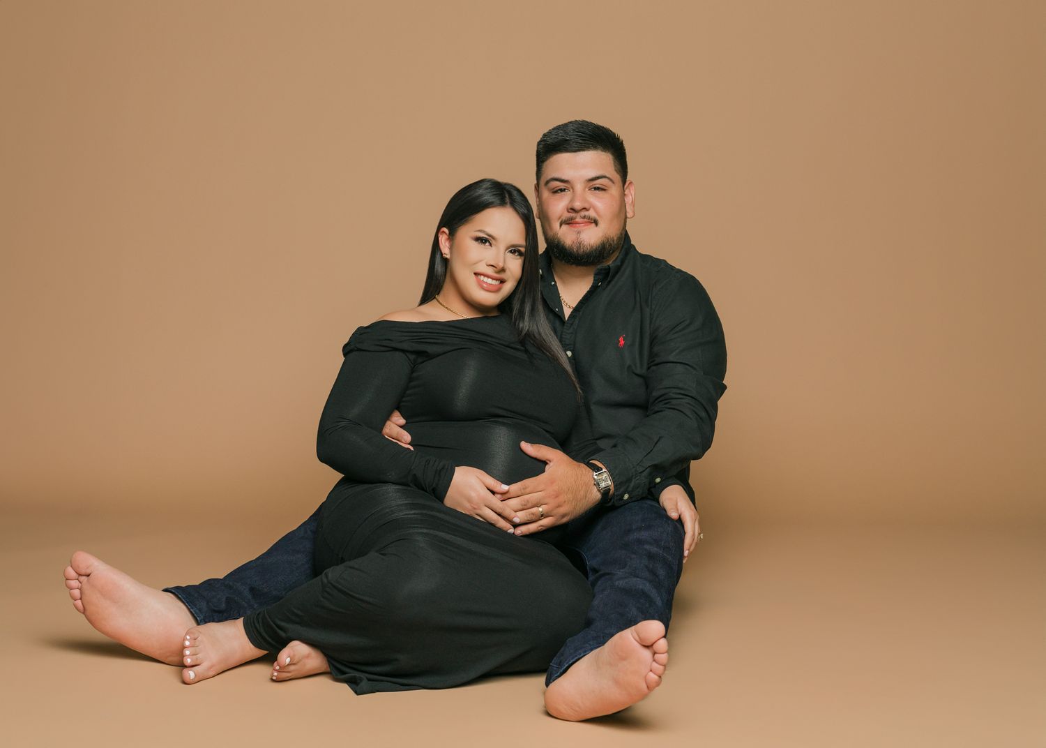 A couple wearing matching black outfits poses together against a warm brown studio backdrop.