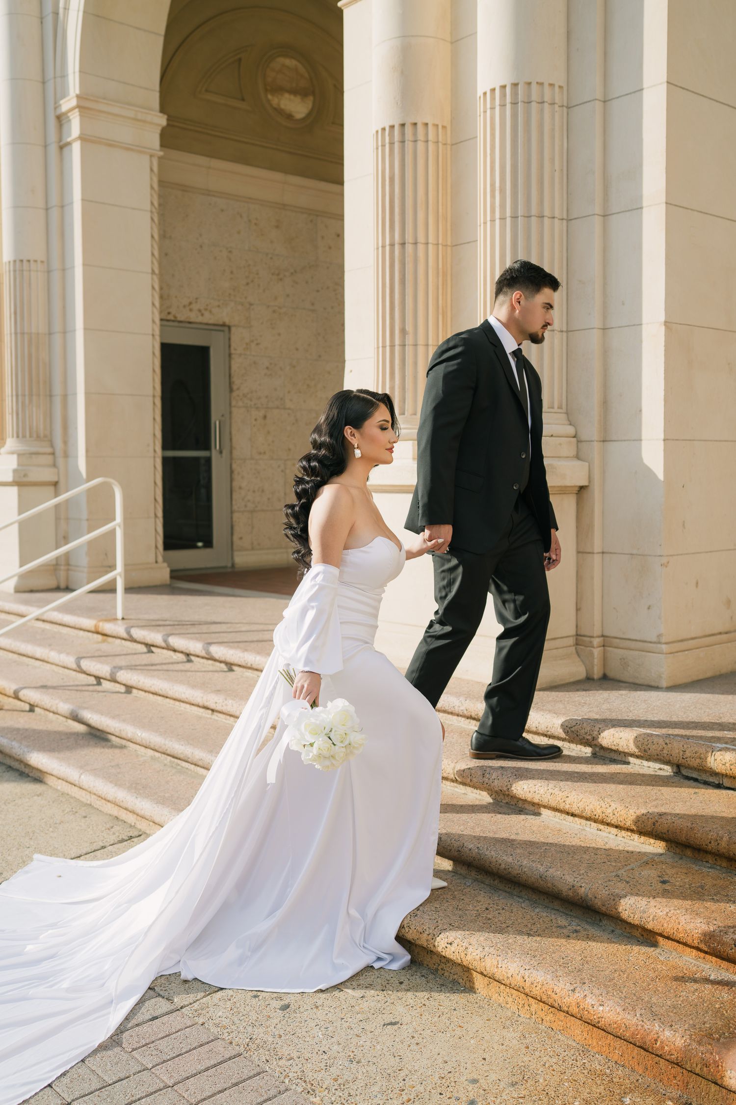 A couple in wedding attire descends elegant stone steps with classical columns in the background.