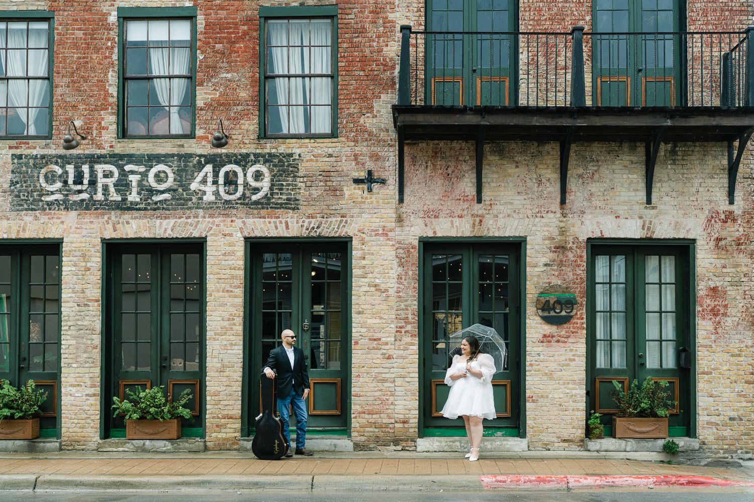 A couple poses separately in front of a rustic brick building with green doors.
