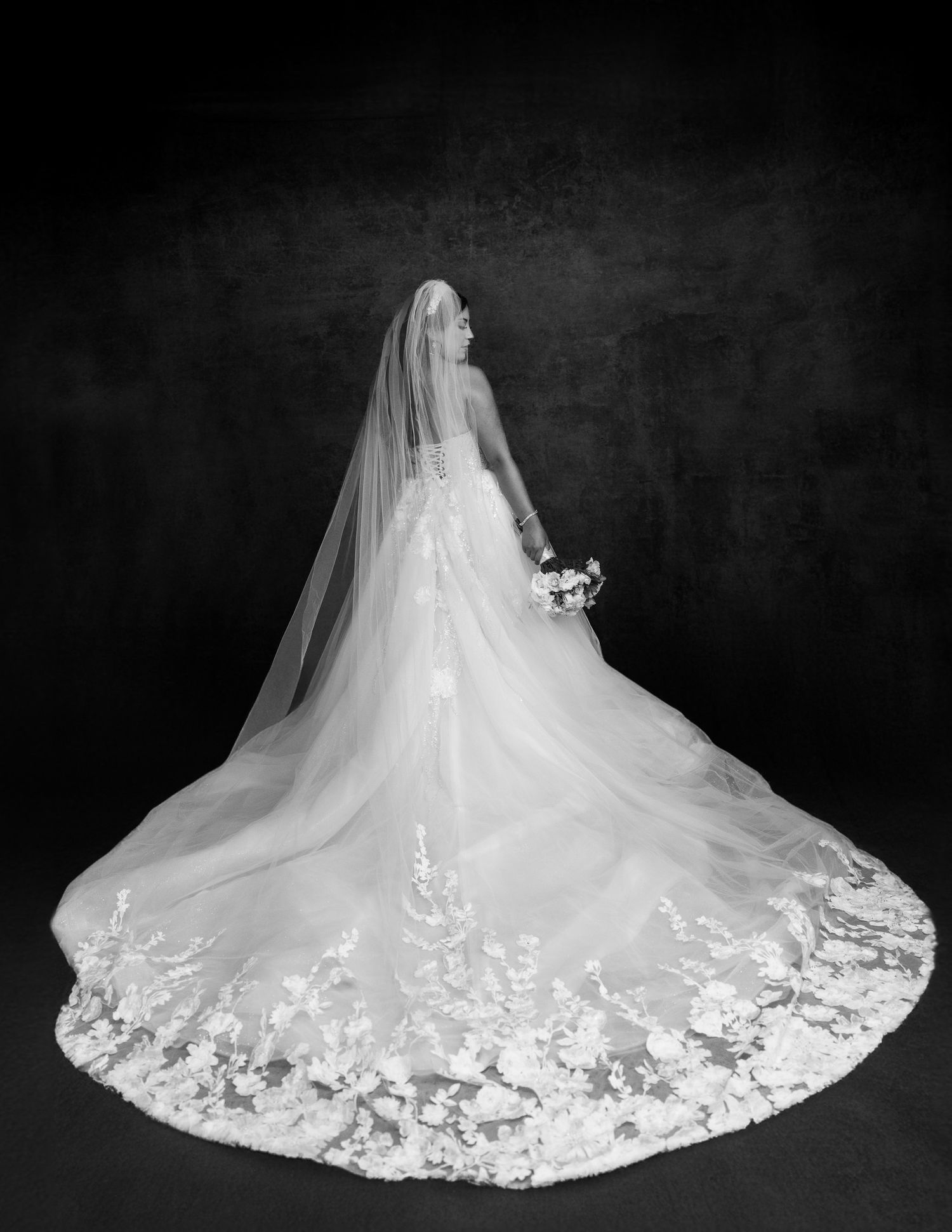 A wedding dress with intricate lace details and long flowing train displayed in dramatic black and white photography.