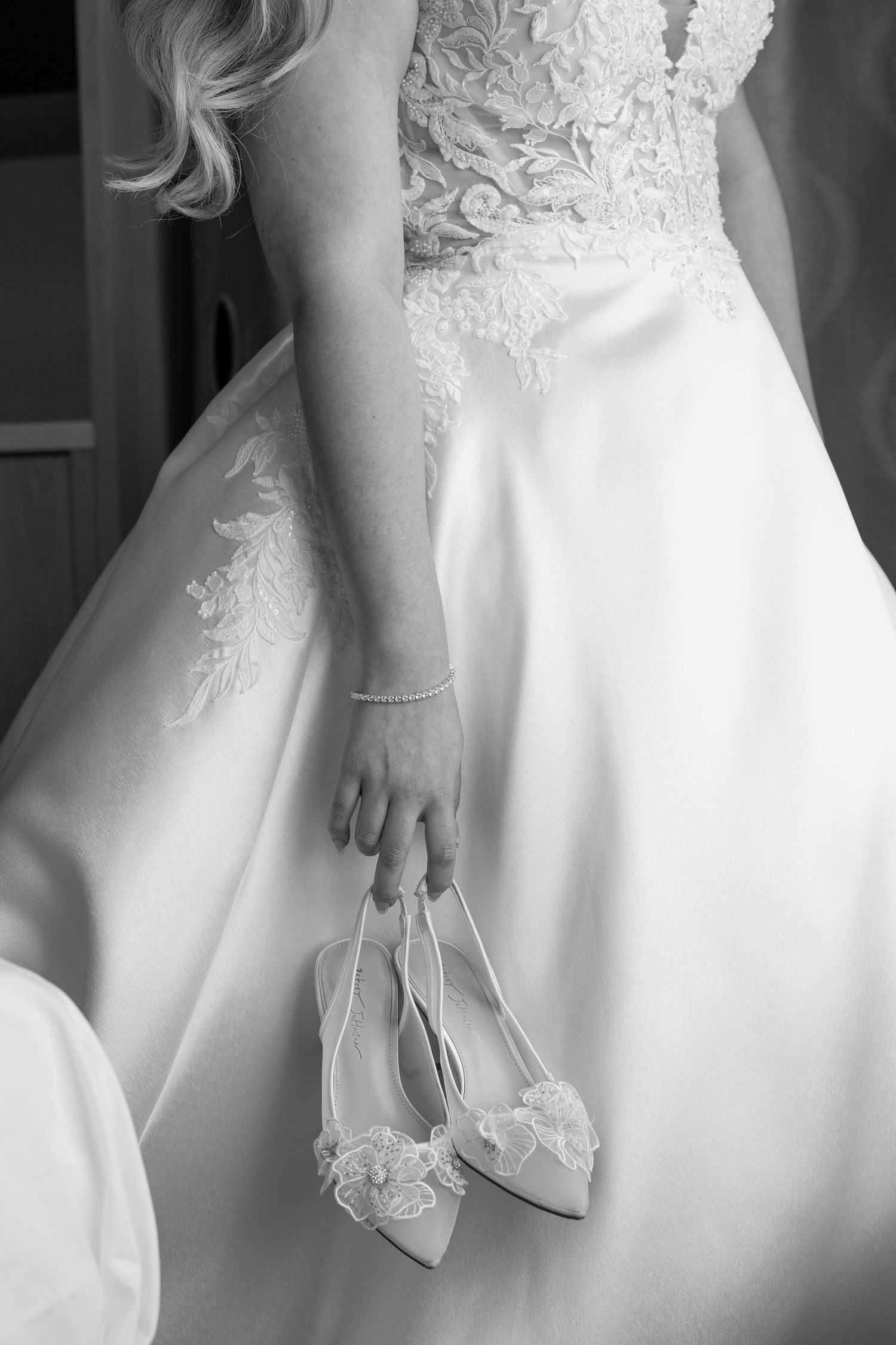Bride holding delicate lace wedding shoes against her white gown in an elegant black and white photo.