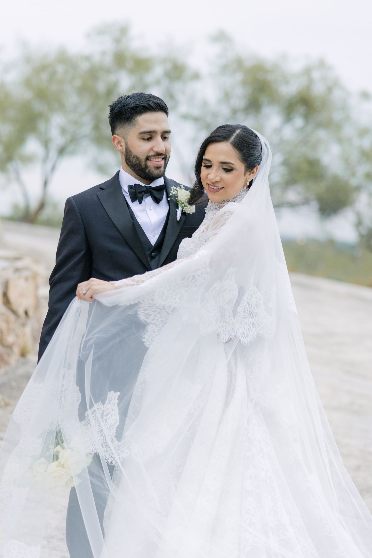 A groom in a black tuxedo holds his bride's flowing lace wedding veil outdoors on their wedding day.