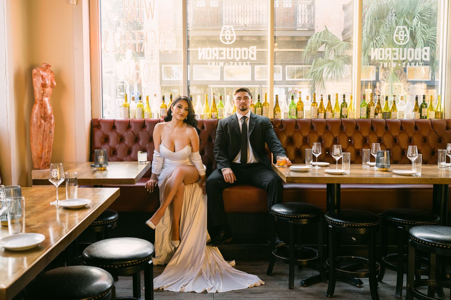 A couple in formal wear sits at an elegant bar with vintage decor and bottle display.