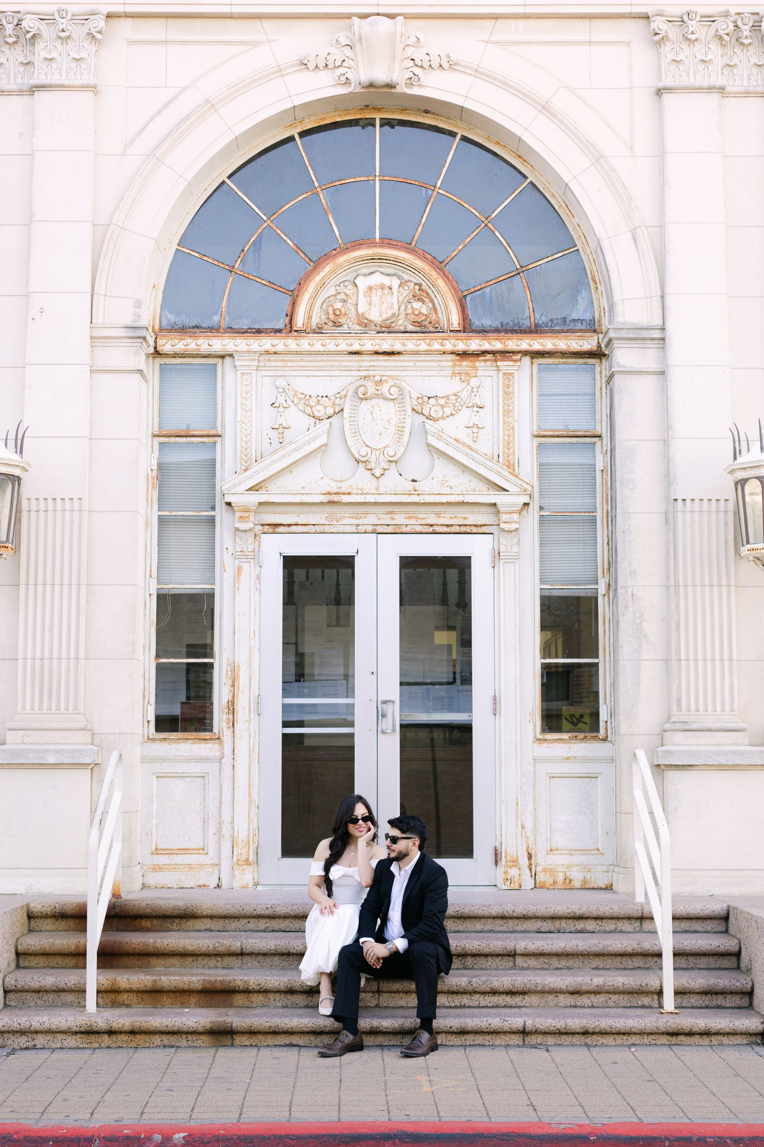 A couple sits on ornate building steps beneath a beautiful arched entrance with decorative details.