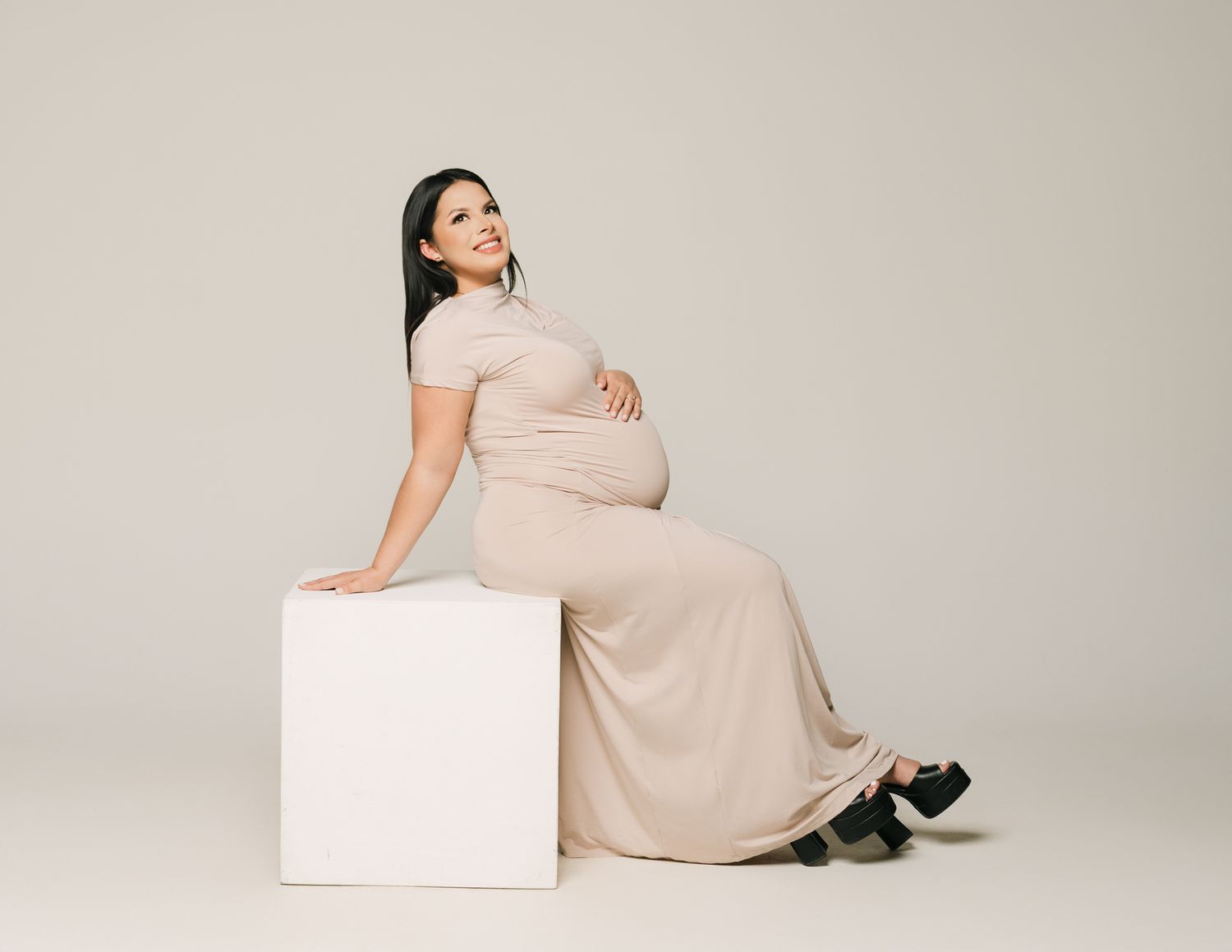 Expectant mother in beige dress sitting gracefully on white cube during studio photoshoot.