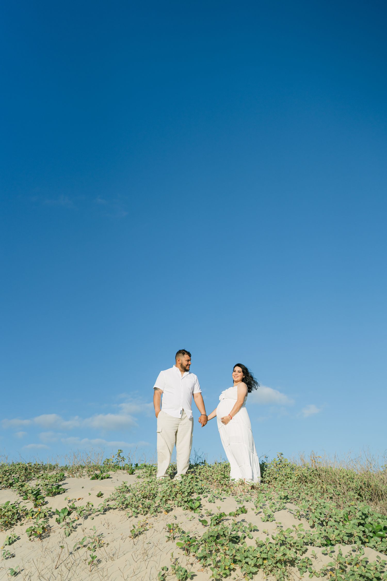 Expectant couple standing together on grassy dune against bright blue sky.