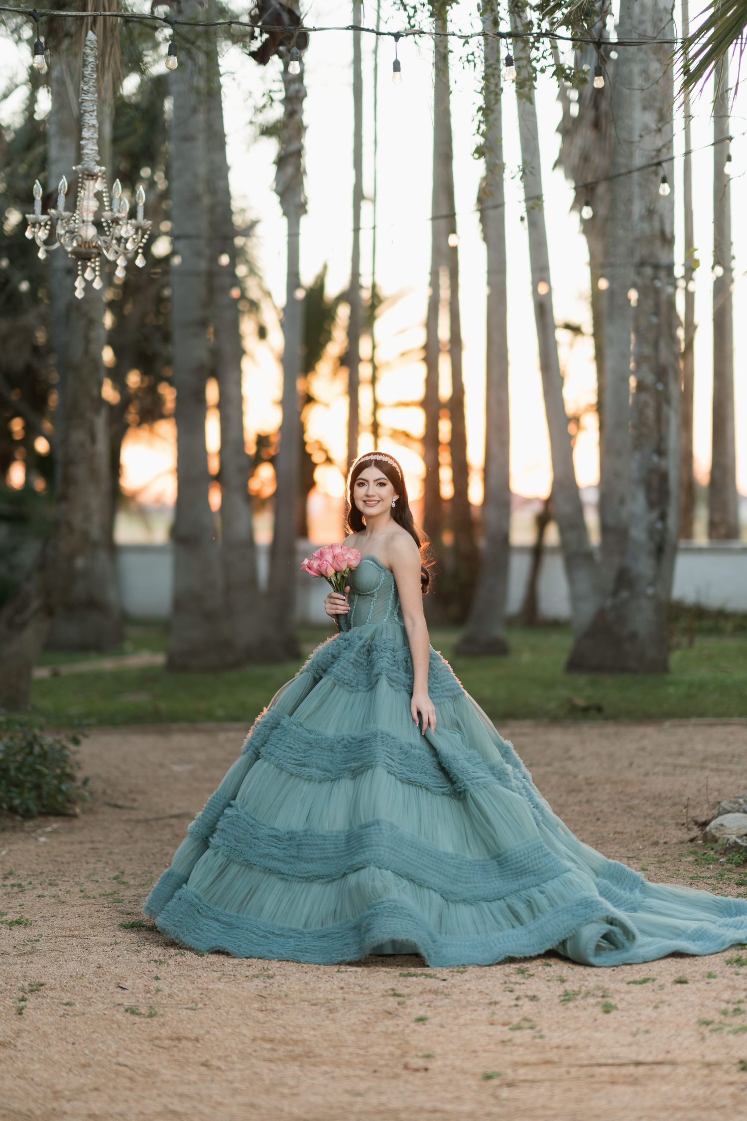 Person in flowing teal gown poses dramatically among tall palm trees at sunset.