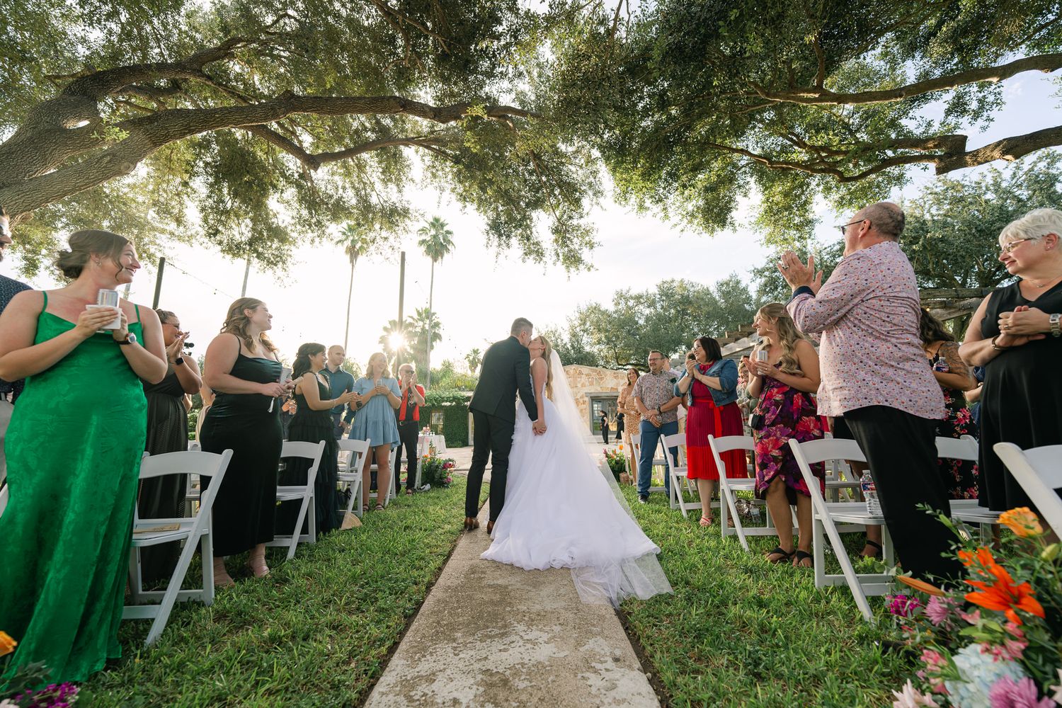 Guests seated on white chairs at an outdoor wedding ceremony under large oak trees.