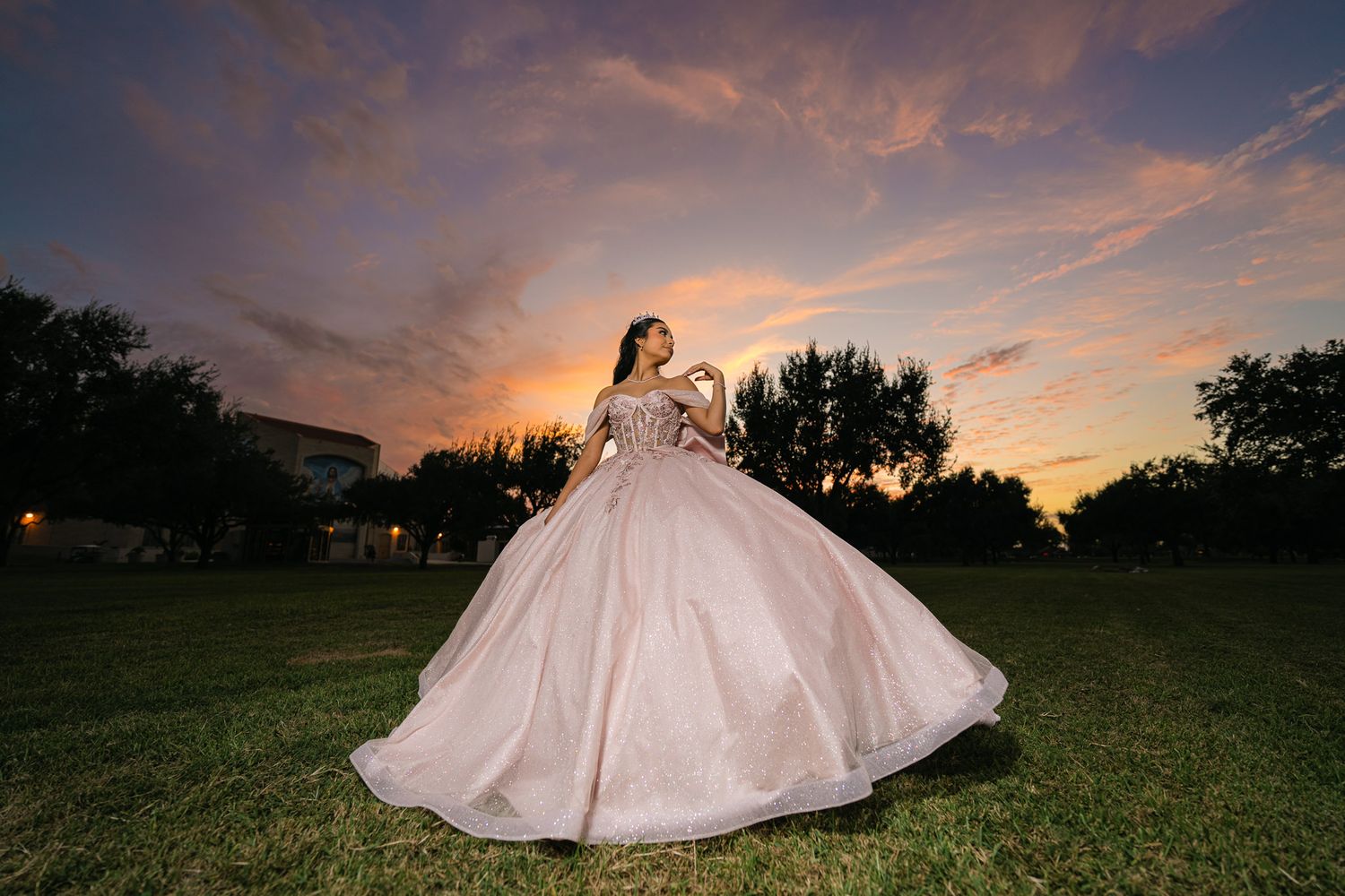 A bride in a voluminous white ballgown twirls on a grassy field at sunset with dramatic orange clouds above.