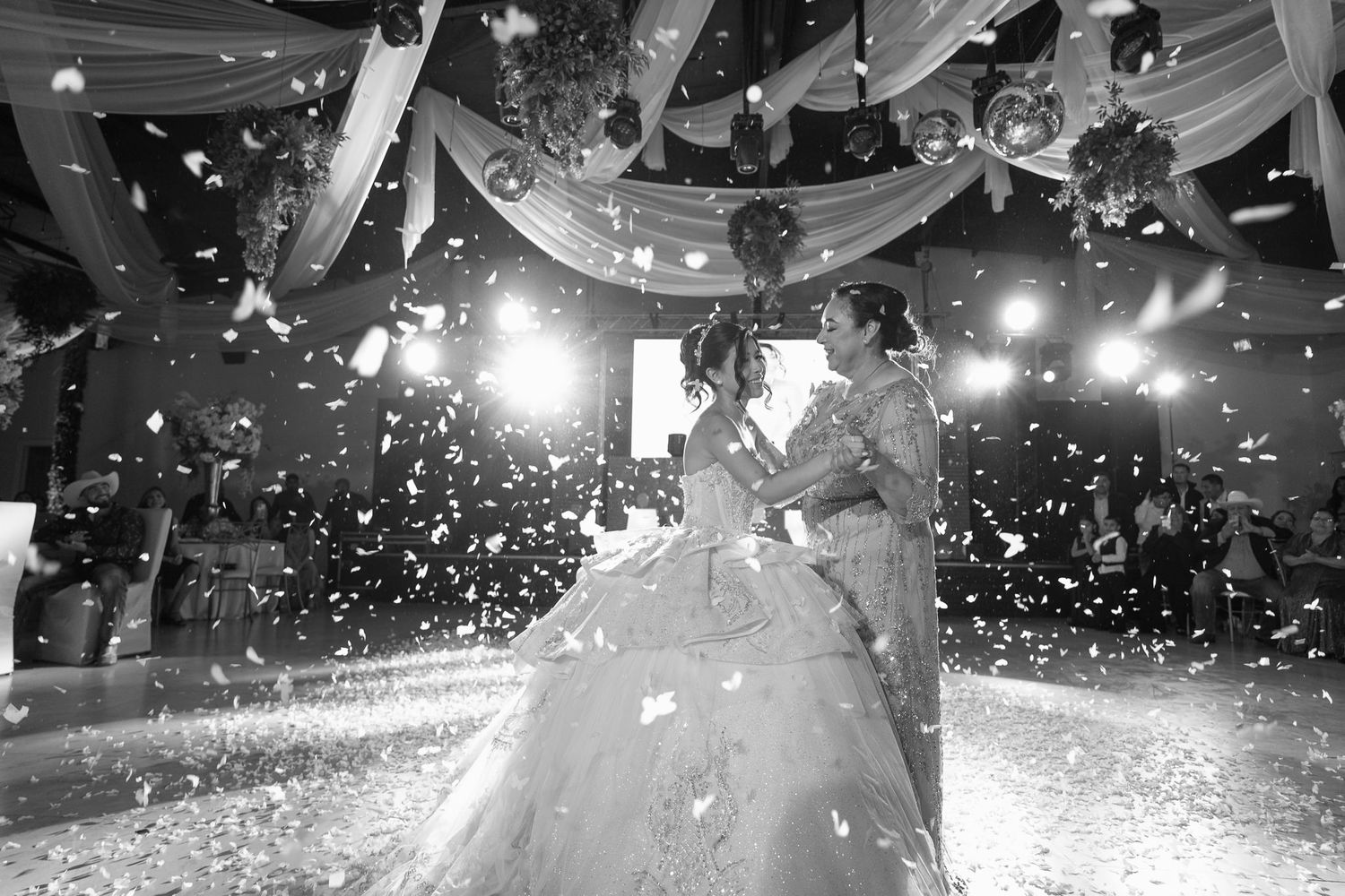 A couple shares their first dance at a wedding reception as confetti falls around them in black and white photography.