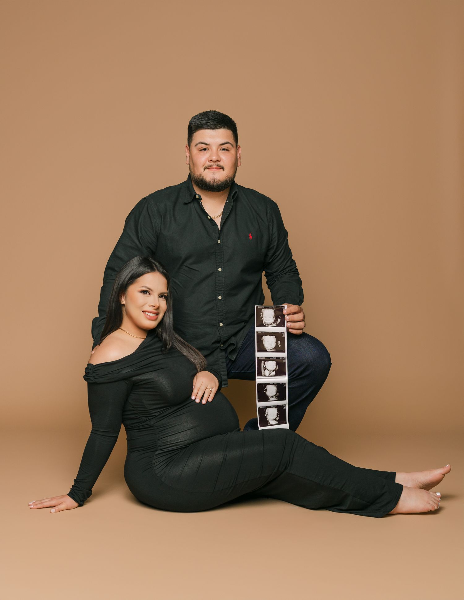 A couple in black attire poses with ultrasound photos against a brown backdrop.