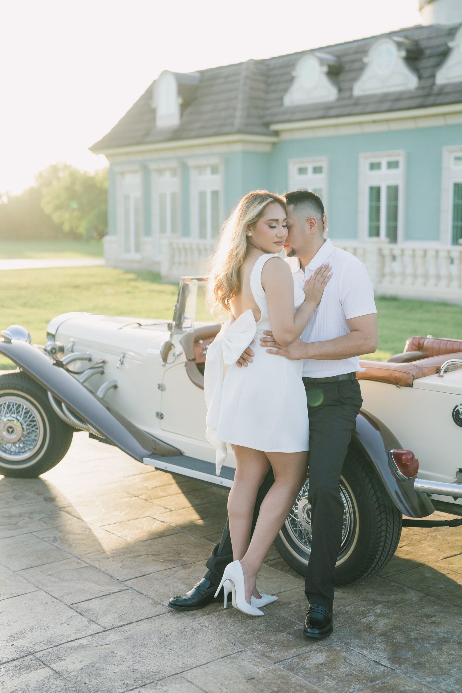 A couple poses romantically against a vintage car with a blue mansion in the background.